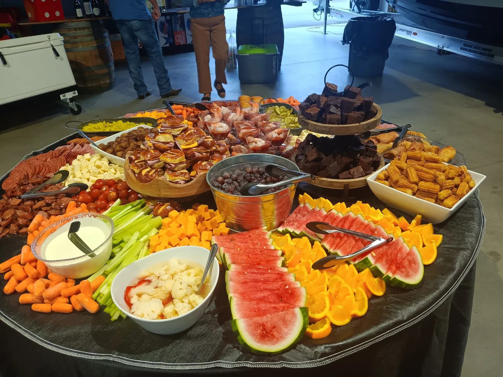 A round grazing table of fruit, meats, cheeses, sliders, shrimp cups, brownies and churros, catered at a boat dealership